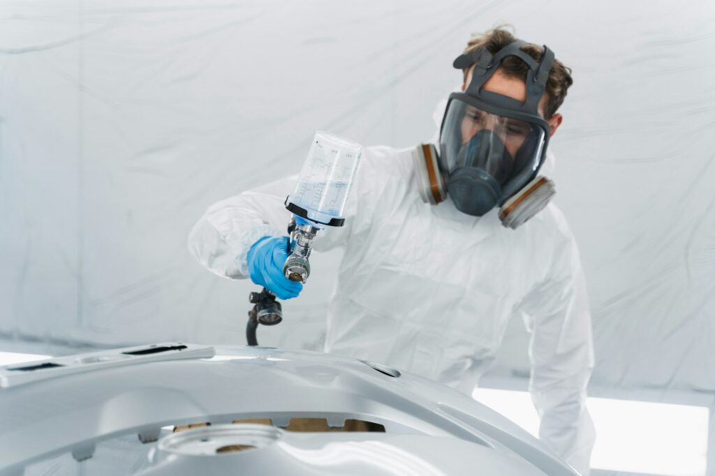 Car painter in a protective suit and mask paint a auto bumper in a painting booth.